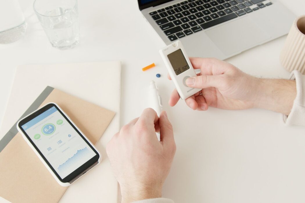 Personne testant sa glycémie avec glucomètre, smartphone et ordinateur portable sur le bureau, avant démo