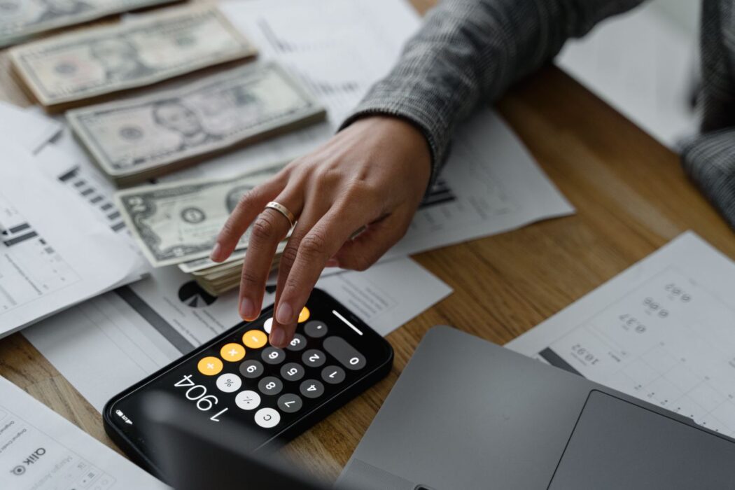 Personne utilisant une calculatrice sur smartphone entourée d'argent et de documents financiers pour gérer son budget